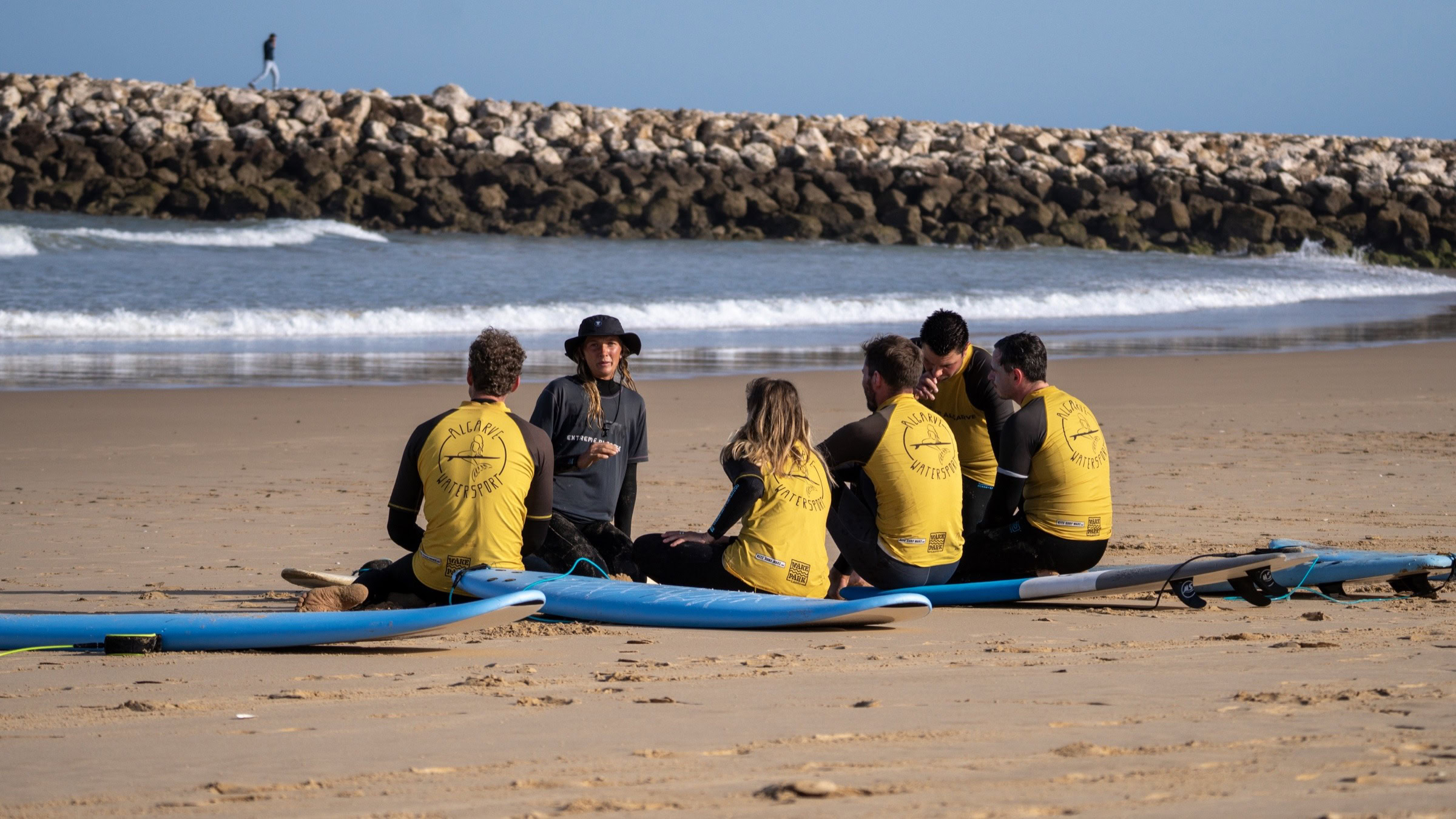 Surf group sitting on Lagos beach with instructor, jetty visible in background, Algarve